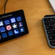 A close-up of a Stream Deck device and part of a keyboard on a wooden desk. The Stream Deck, with 15 programmable buttons, displays various icons and labels, including "Home," "Someday," "Saturday," "Tomorrow," and "Complete." The keyboard, partially visible, features standard keys, including "esc," "tab," "caps lock," and "shift." The scene suggests a workspace setup for productivity or content creation.