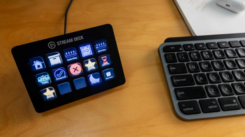 A close-up of a Stream Deck device and part of a keyboard on a wooden desk. The Stream Deck, with 15 programmable buttons, displays various icons and labels, including "Home," "Someday," "Saturday," "Tomorrow," and "Complete." The keyboard, partially visible, features standard keys, including "esc," "tab," "caps lock," and "shift." The scene suggests a workspace setup for productivity or content creation.