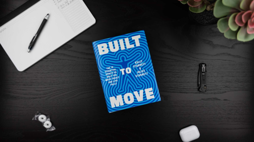 A flat-lay image of a desk featuring the book "Built to Move" by Kelly Starrett and Juliet Starrett, with a bold blue cover and white text. Nearby are a black pen on an open notebook, two wrapped mints, a folded pocket knife, a pair of white AirPods, and a succulent plant in the corner. The desk surface is dark and textured.