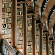 A grand library interior with tall, ornate wooden bookshelves filled with neatly arranged books. The shelves are framed by decorative columns, and the ceiling features a series of curved wooden arches with metal accents, creating a symmetrical and majestic architectural design. Warm lighting enhances the richness of the wood and the historic ambiance of the space.