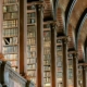 A grand library interior with tall, ornate wooden bookshelves filled with neatly arranged books. The shelves are framed by decorative columns, and the ceiling features a series of curved wooden arches with metal accents, creating a symmetrical and majestic architectural design. Warm lighting enhances the richness of the wood and the historic ambiance of the space.