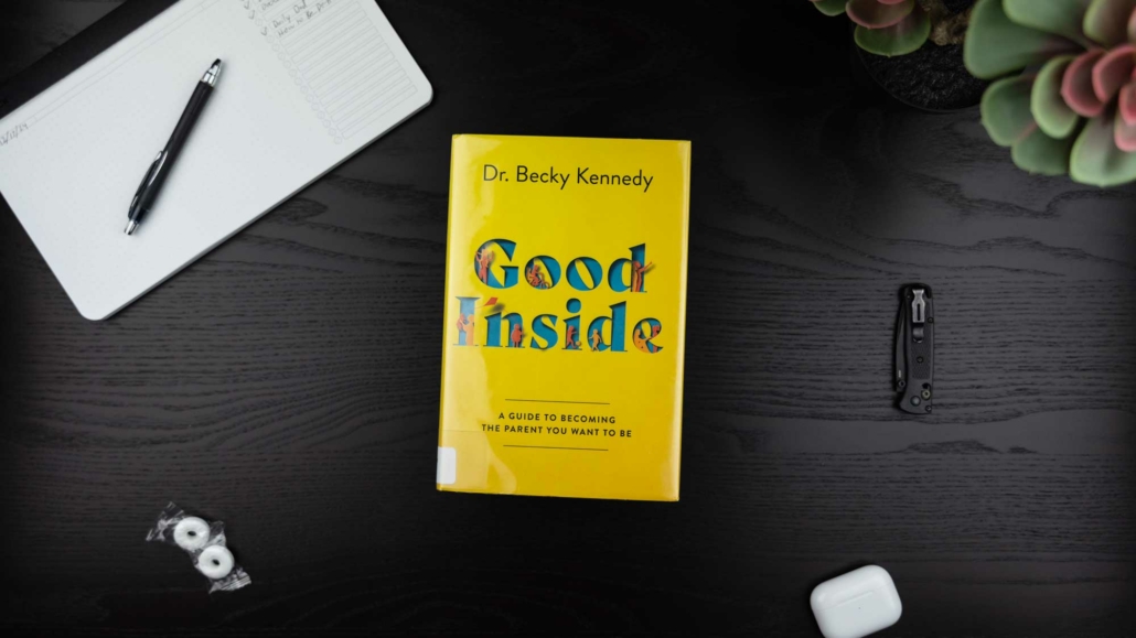 A flat-lay image of a desk featuring the book "Good Inside" by Dr. Becky Kennedy, with a bright yellow cover and colorful cutout-style text. The subtitle reads, "A Guide to Becoming the Parent You Want to Be." Surrounding items include a black pen on an open notebook, two wrapped mints, a folded pocket knife, white AirPods, and a succulent plant in the corner. The desk surface is dark wood.