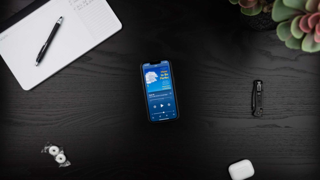 A flat-lay image of a desk featuring a smartphone displaying the audiobook "How to Be Perfect" by Michael Schur on its screen. Surrounding the phone are a black pen on an open notebook, two wrapped mints, a folded pocket knife, white AirPods, and a succulent plant in the corner. The desk surface is dark wood with a clean, organized layout.