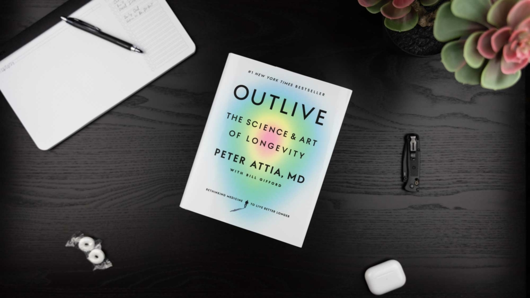 A flat-lay image of a desk featuring the book "Outlive: The Science & Art of Longevity" by Peter Attia, MD, with Bill Gifford. The cover is white with a subtle gradient design. Surrounding items include a black pen on an open notebook, two wrapped mints, a folded pocket knife, white AirPods, and a succulent plant in the corner. The desk surface is dark wood and neatly organized.