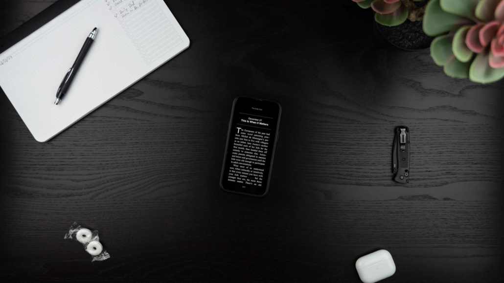 A flat-lay image of a desk featuring a smartphone displaying a text passage titled "This Is When It Matters" from "The Daily Dad." The text is white on a black background. Surrounding items include a black pen on an open notebook, two wrapped mints, a folded pocket knife, white AirPods, and a succulent plant in the corner. The desk surface is dark wood with a minimalist and organized arrangement.