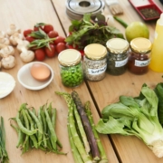 A wooden table filled with fresh ingredients, including leafy greens, asparagus, green beans, chives, mushrooms, cherry tomatoes, and herbs. Small bowls contain cheese, cream, and an egg. Jars of spices and seeds labeled "Sunflower," "Pumpkin," and "Paprika" are placed nearby. A yellow squeeze bottle, a green apple, and kitchen utensils are also present, suggesting a cooking or meal preparation setting.