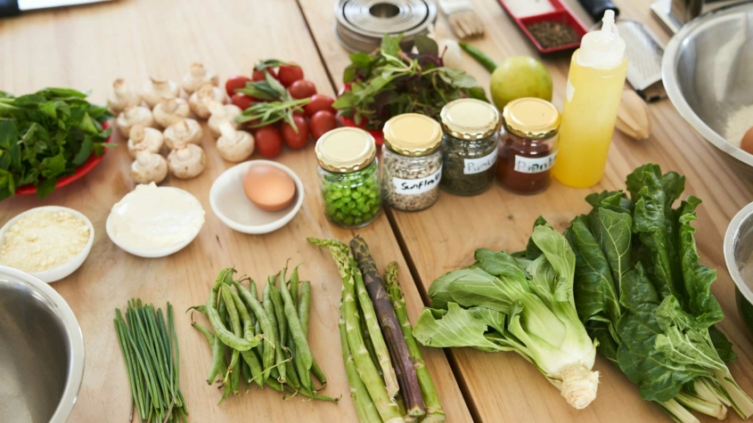 A wooden table filled with fresh ingredients, including leafy greens, asparagus, green beans, chives, mushrooms, cherry tomatoes, and herbs. Small bowls contain cheese, cream, and an egg. Jars of spices and seeds labeled "Sunflower," "Pumpkin," and "Paprika" are placed nearby. A yellow squeeze bottle, a green apple, and kitchen utensils are also present, suggesting a cooking or meal preparation setting.